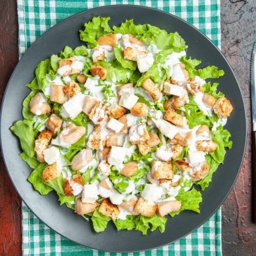 top-view-caesar-salad-oval-plate-green-white-checkered-tablecloth-fork-knife-dark-red-background